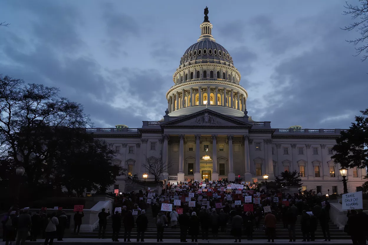 Protestors holding signs fill the steps of the illuminated U.S. Capitol building at dusk.