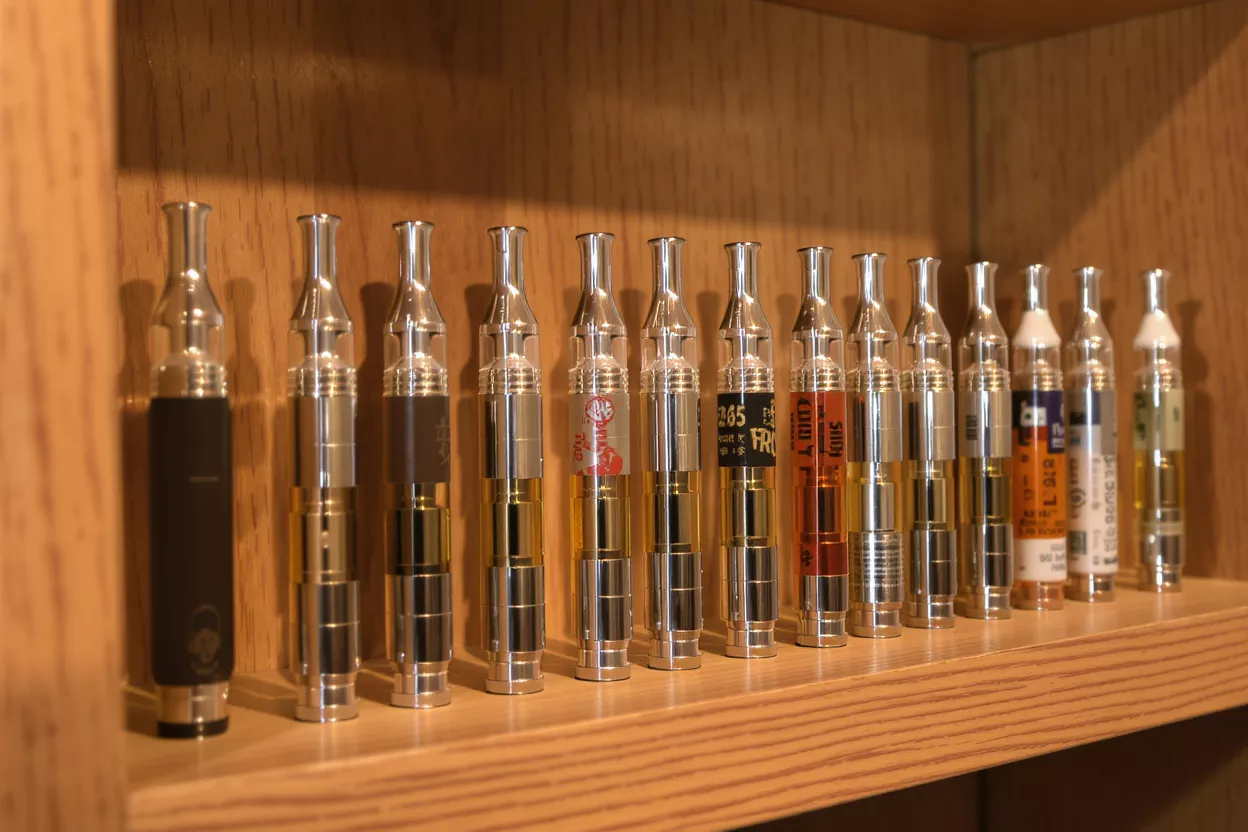 A row of various vape cartridges and one vape pen on a wooden shelf.