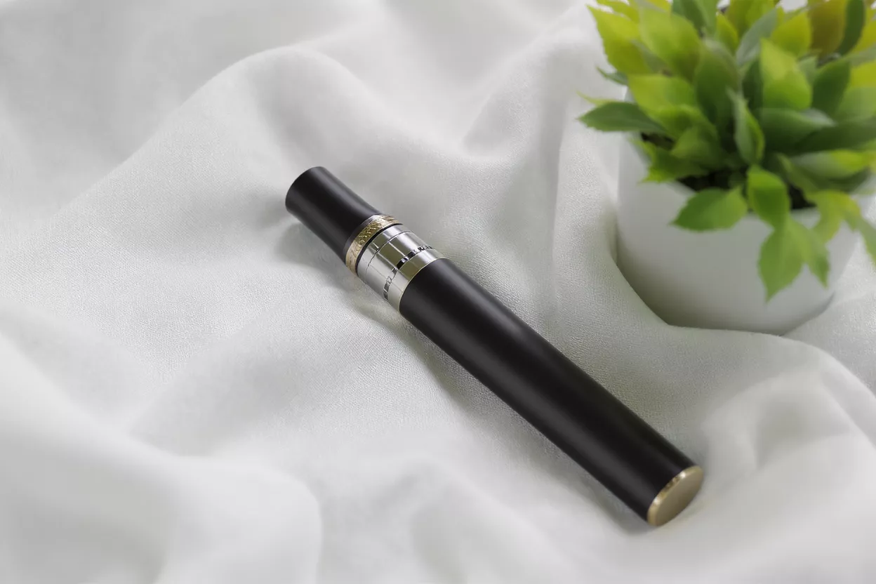 Black vape pen with gold and silver accents on white fabric next to a plant.