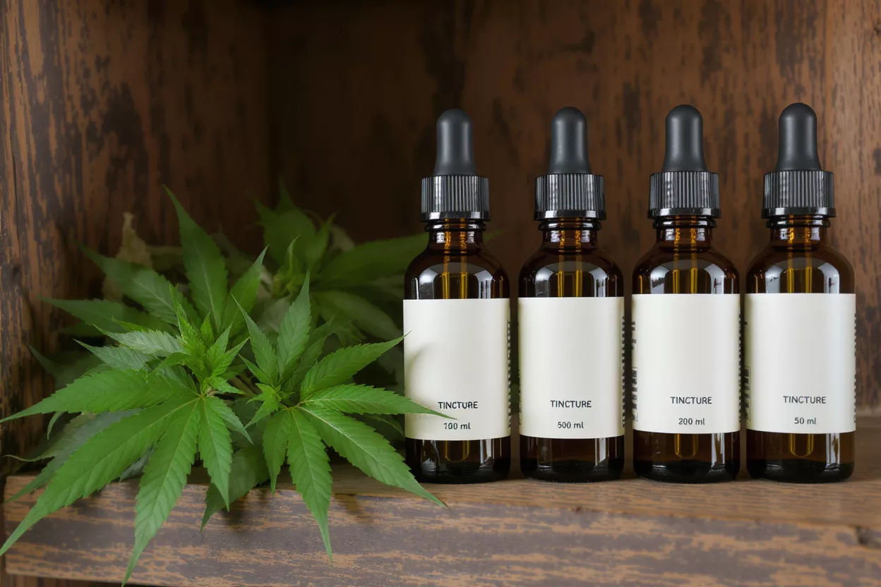 Four amber tincture bottles with white labels next to fresh cannabis leaves on a wooden shelf.