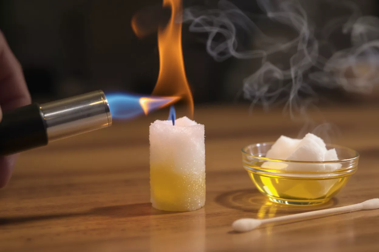 A hand holding a torch lighting a sugar candle, with a bowl of oil and sugar nearby.