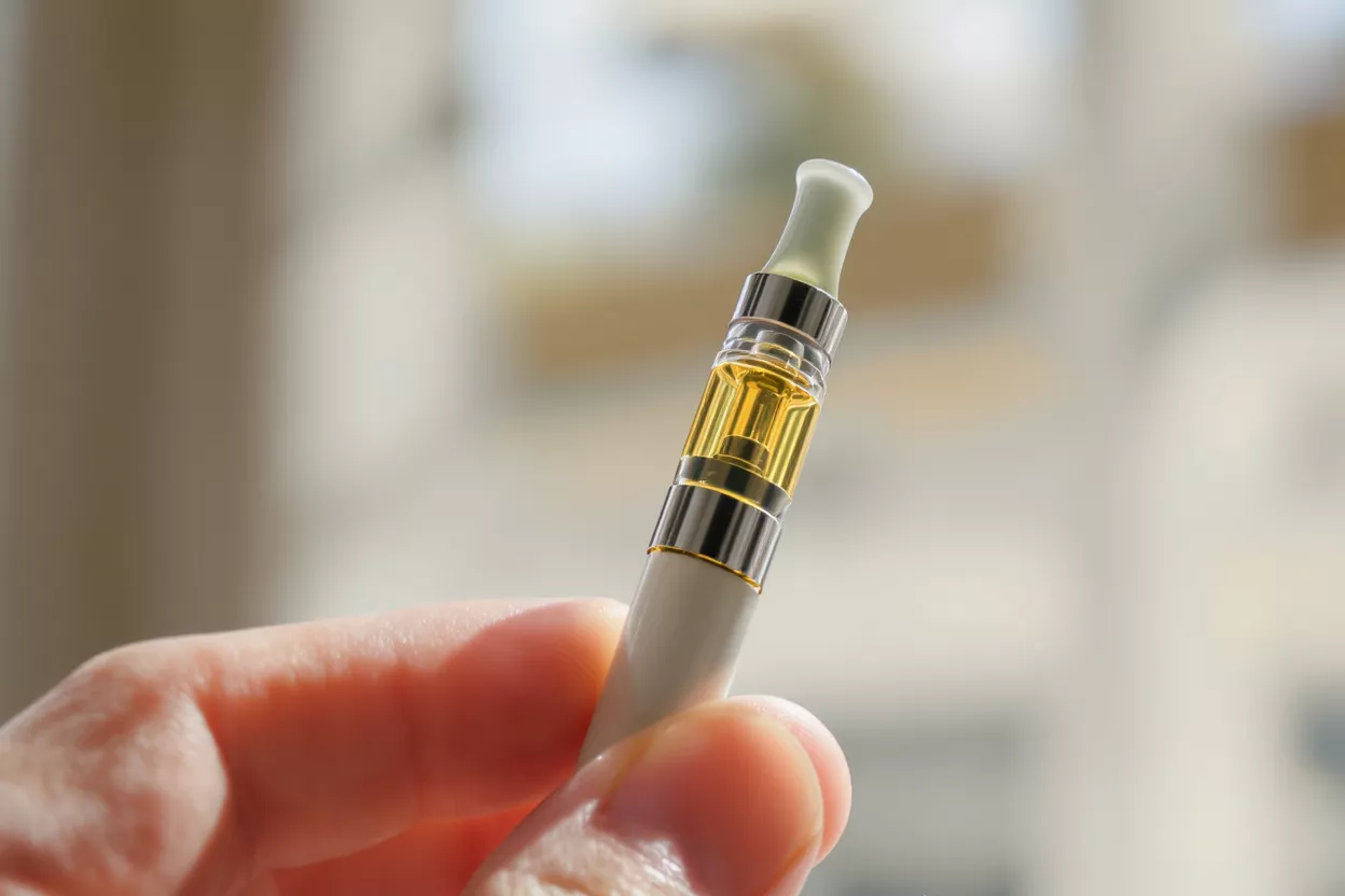 A hand holds a vape cartridge filled with golden oil against a blurred background.