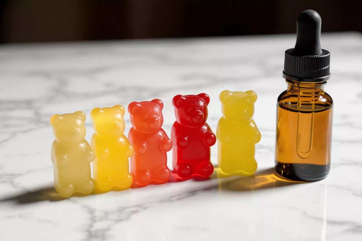 Five colorful gummy bears next to a CBD oil dropper bottle on a marble surface.