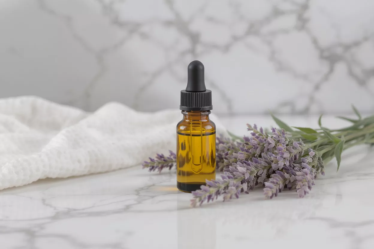 Amber dropper bottle with lavender essential oil, white towel, and lavender sprigs on marble.