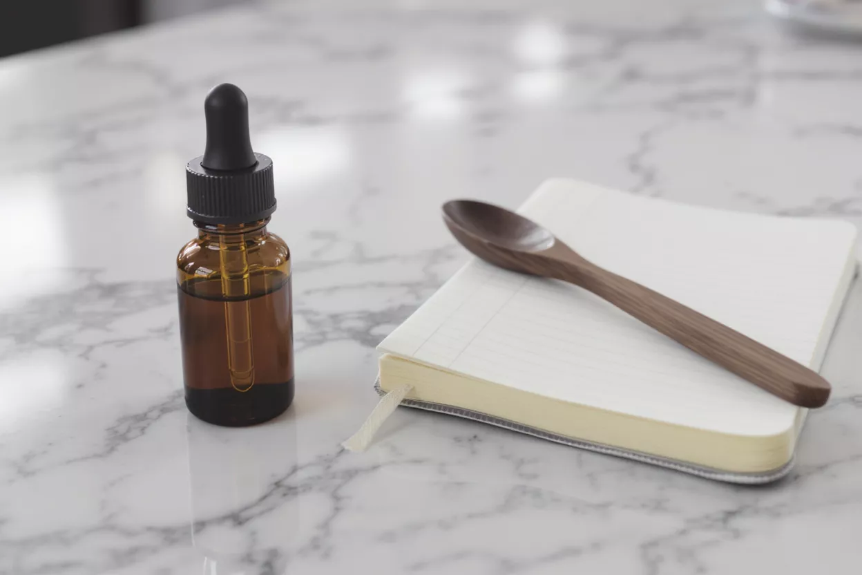 Amber dropper bottle, wooden spoon, and open notebook on a marble surface.