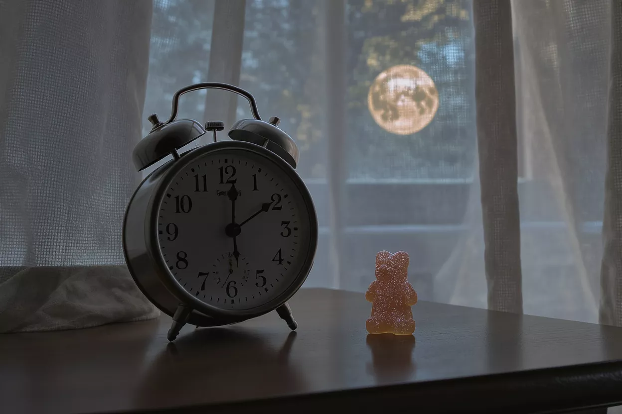 Réveil et Bonbon Ourson Un réveil et un bonbon ourson sur une table avec une pleine lune dehors.
