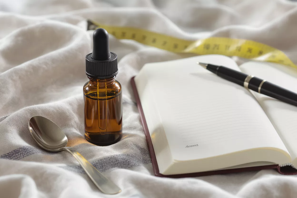 Amber dropper bottle, spoon, open notebook, pen, and measuring tape on fabric.