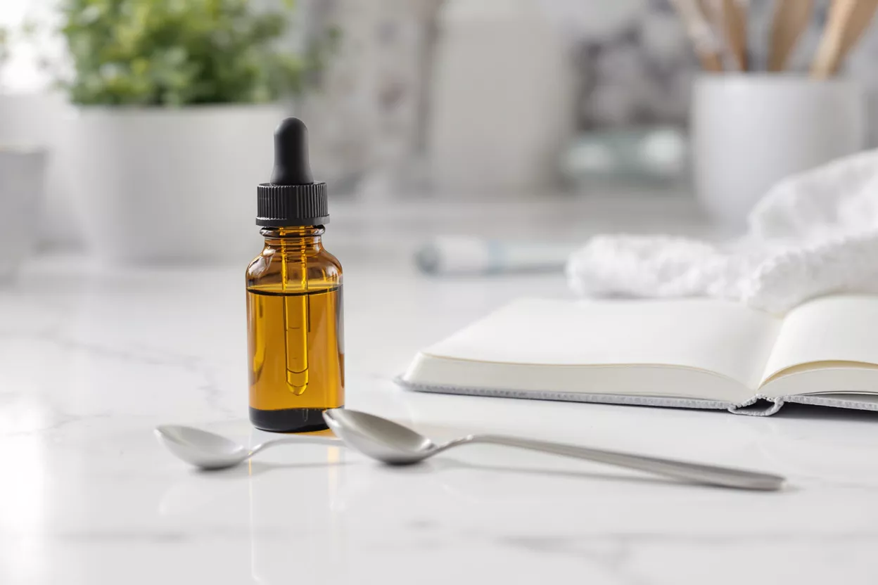 Amber Dropper Bottle with Spoons and Notebook Amber dropper bottle with liquid, two spoons, and an open notebook on a marble surface.