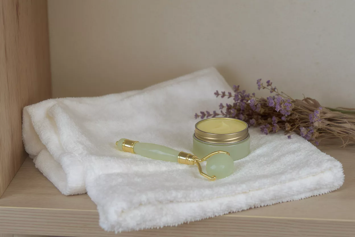 Skincare set with jade roller, cream, and lavender on a white towel.