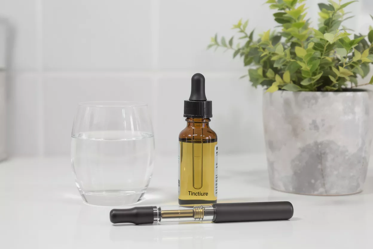 Amber tincture bottle, vape pen, water glass, and plant on white surface.