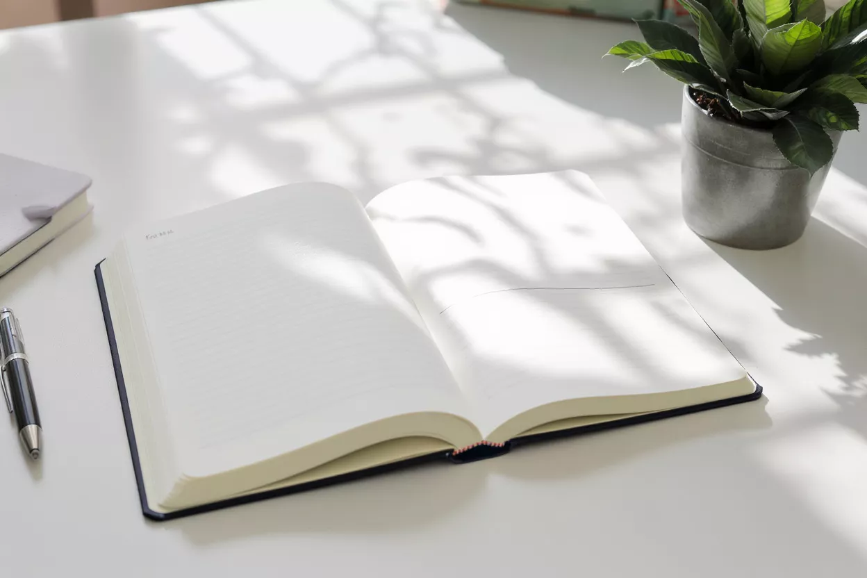 An open notebook with blank pages, a pen, and a plant on a white desk.