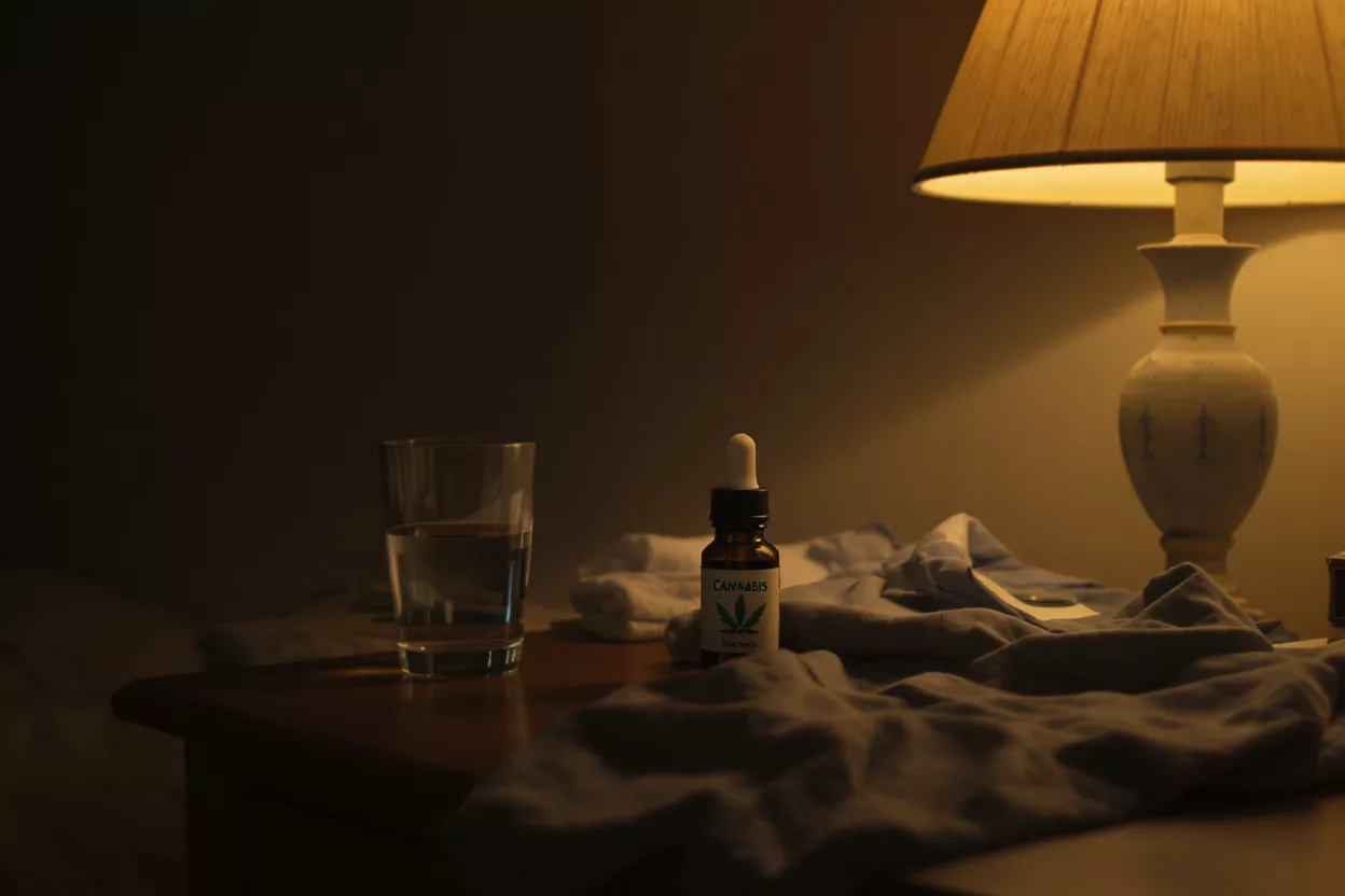 Cannabis oil bottle, water glass, and lamp on a dark bedside table.