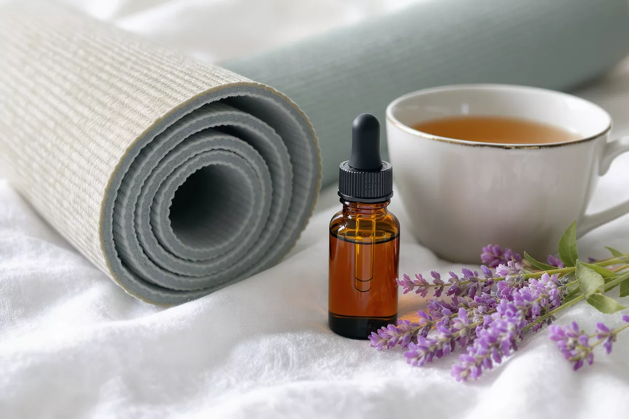 Yoga mat, essential oil bottle, lavender, and tea cup on white fabric.