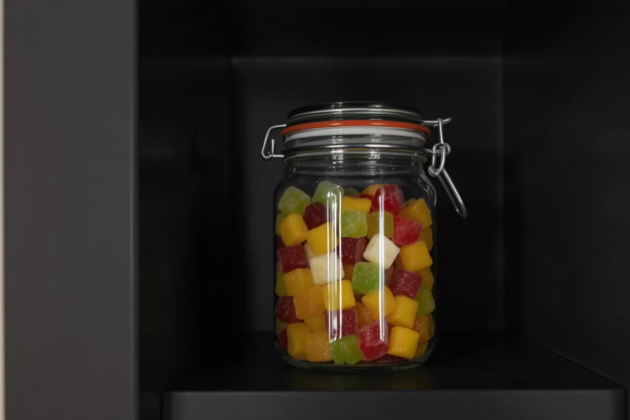 A glass jar filled with colorful square fruit gummies sits on a dark shelf.