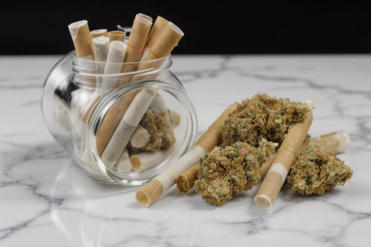 Cannabis buds and pre-rolled joints in a glass jar on a marble surface.