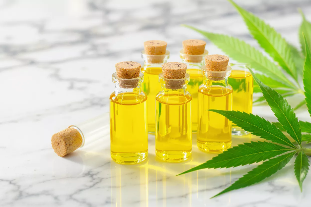 Small glass bottles of golden CBD oil with cannabis leaves on marble surface.