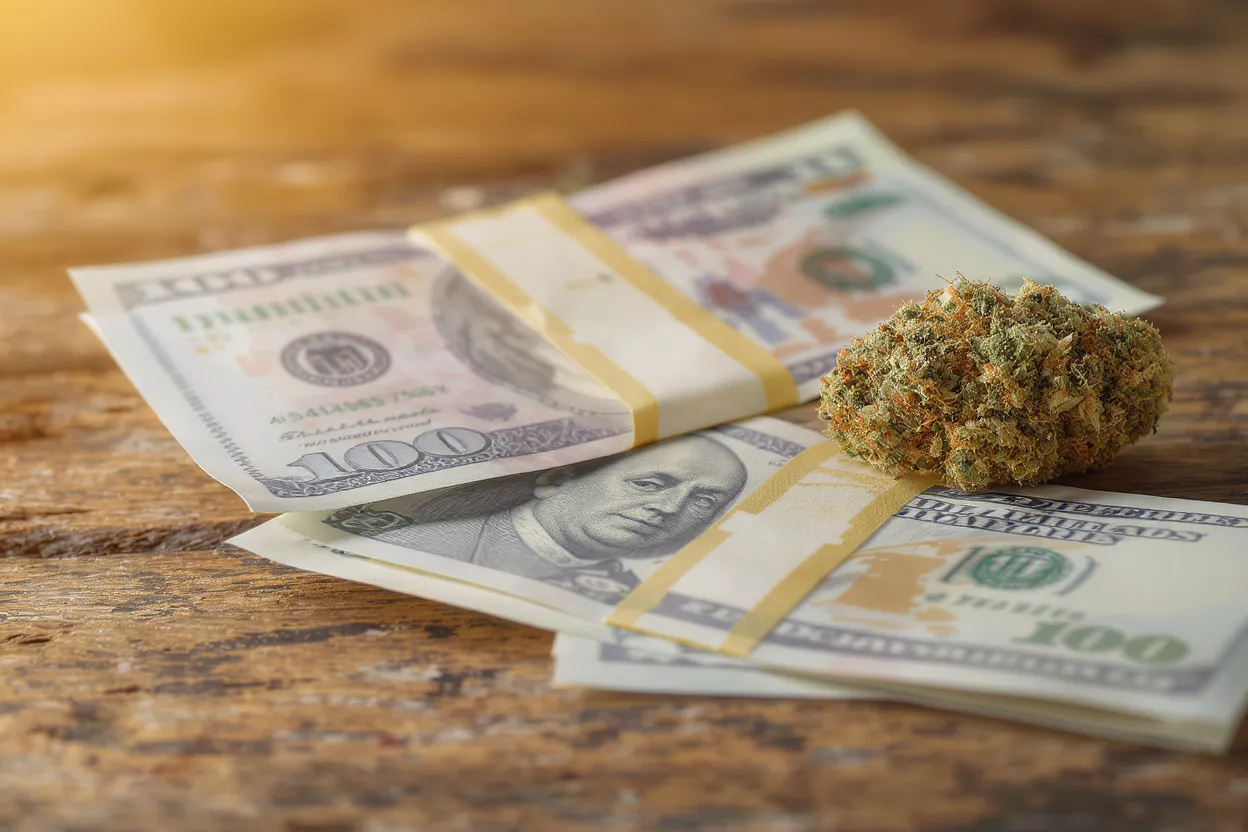 A cannabis bud resting on stacks of hundred-dollar bills on a wooden surface.