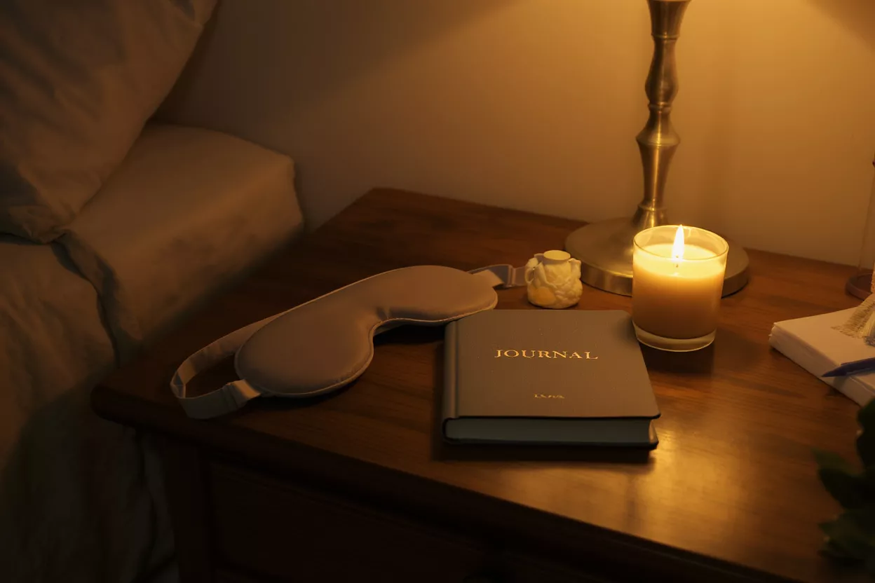 Sleep mask, journal, and lit candle on a wooden bedside table.