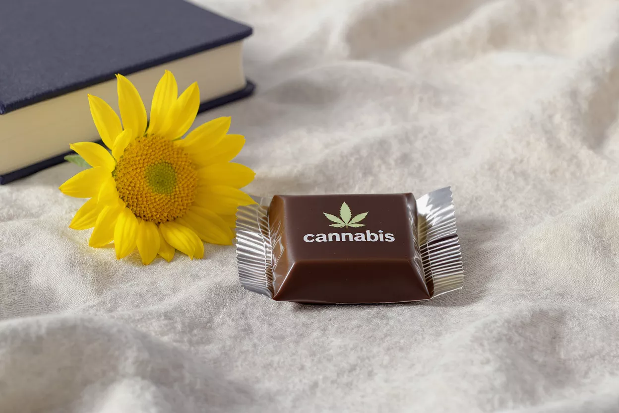Chocolate square with cannabis logo, sunflower, and book on a cream fabric.