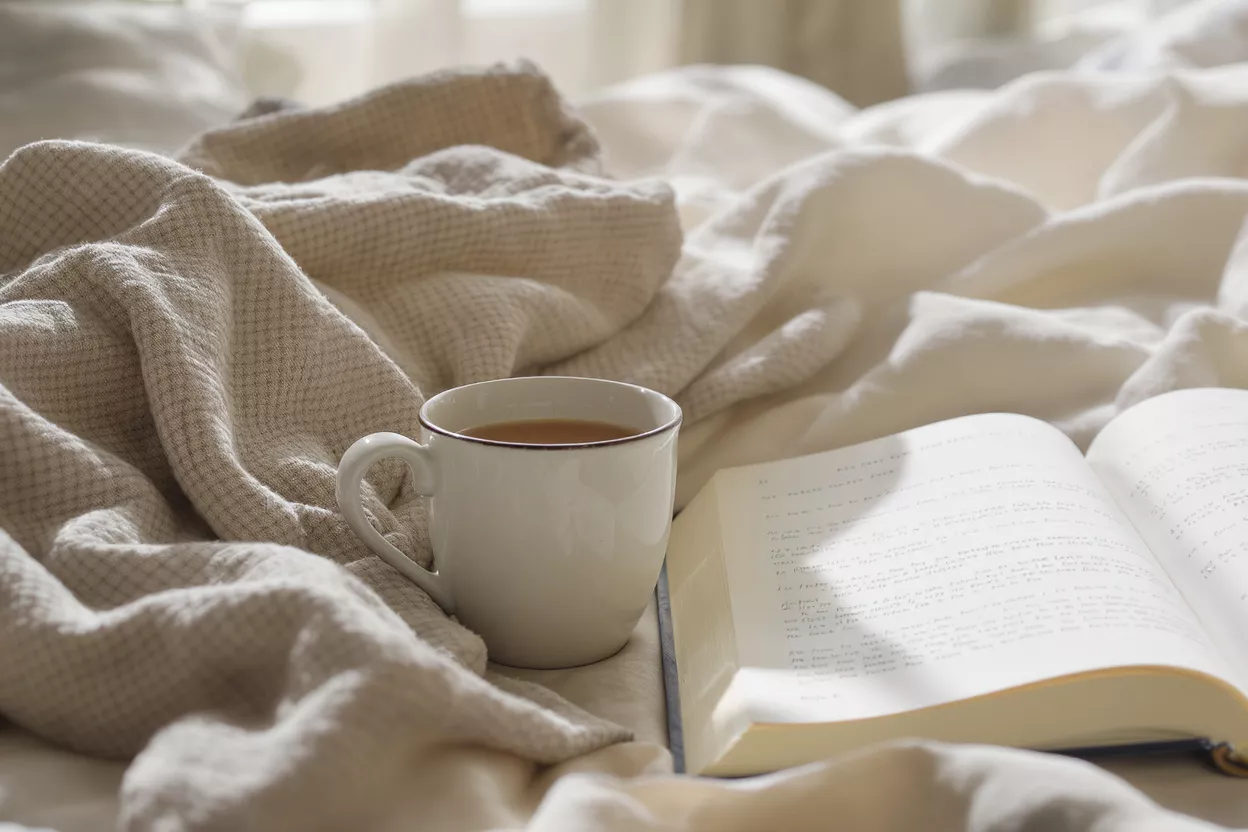 Café et livre pour un matin cosy Une tasse blanche de café et un livre ouvert sur un lit avec des couvertures douces.