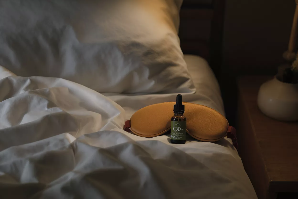 Botella de aceite de CBD y antifaz para dormir sobre ropa de cama blanca para un descanso reparador.