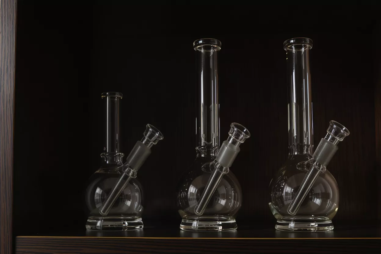 Three clear glass water pipes with round bases and straight necks on a dark shelf.