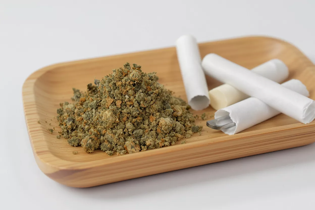 Herbal Mixture and Rolling Accessories A wooden tray with a pile of green and brown herbal mixture and white rolling papers.