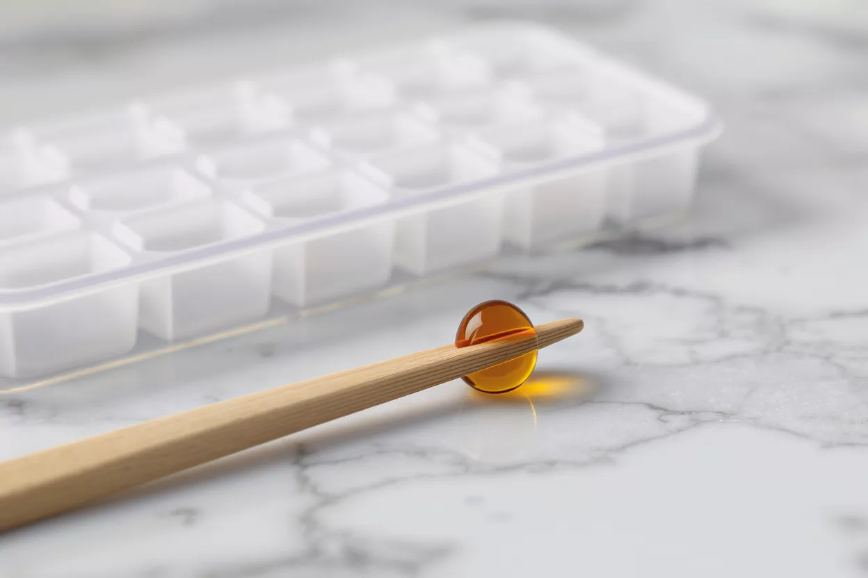 An amber-colored lozenge or capsule balanced on a wooden stick on a marble surface.
