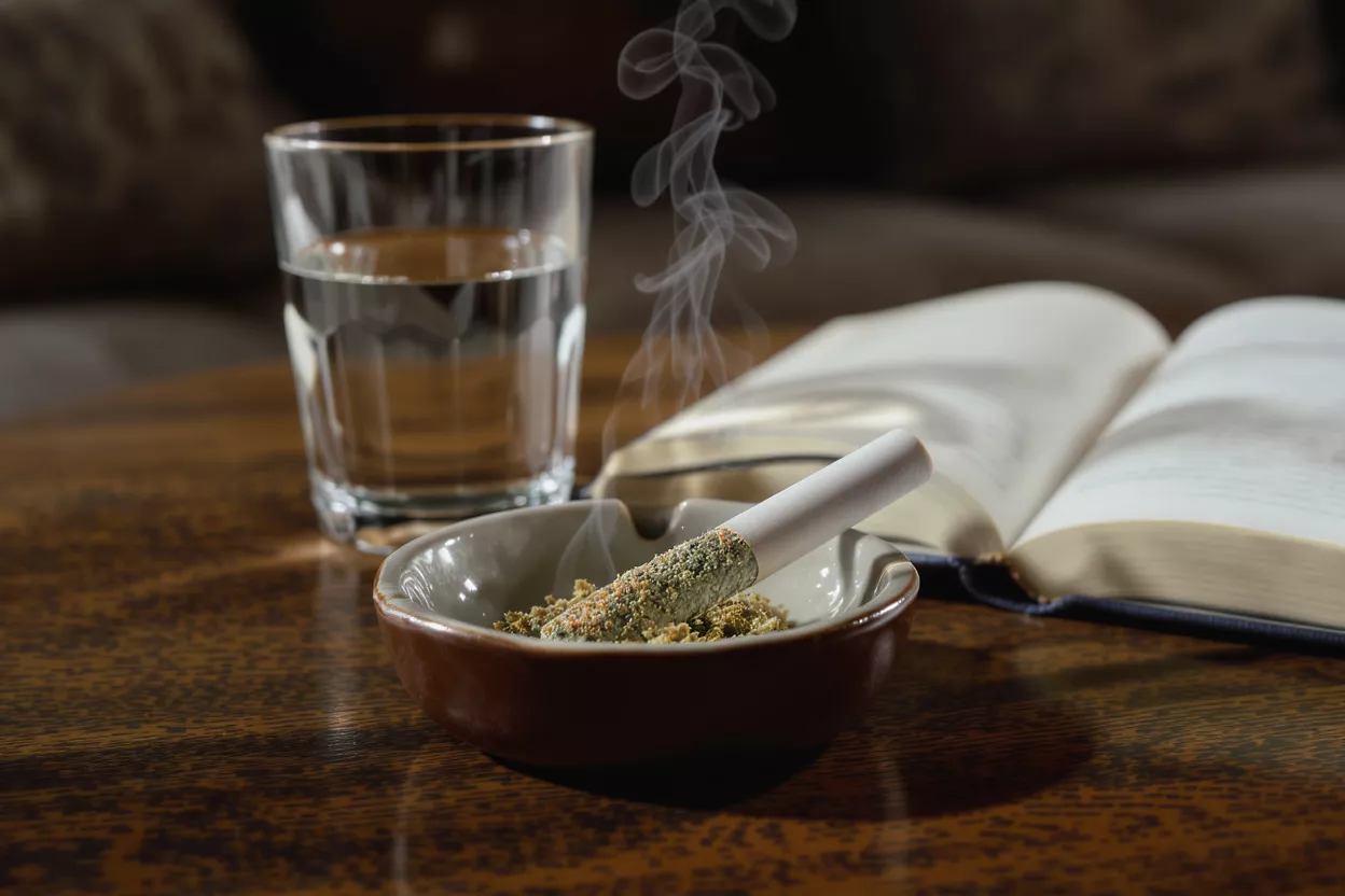 Smoking Joint While Reading A lit joint in an ashtray, glass of water, and open book on a wooden table.