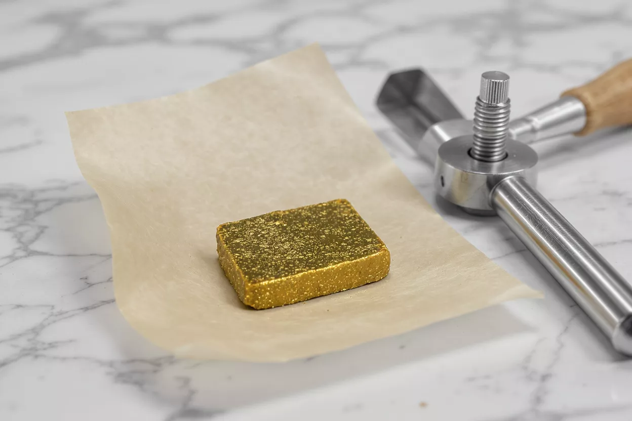 A golden chocolate bar on parchment paper next to a metal press.