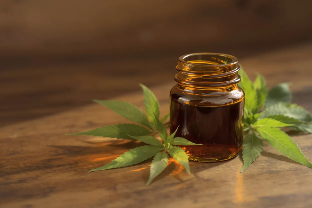 Amber glass bottle of CBD oil with green hemp leaves on a wooden surface.