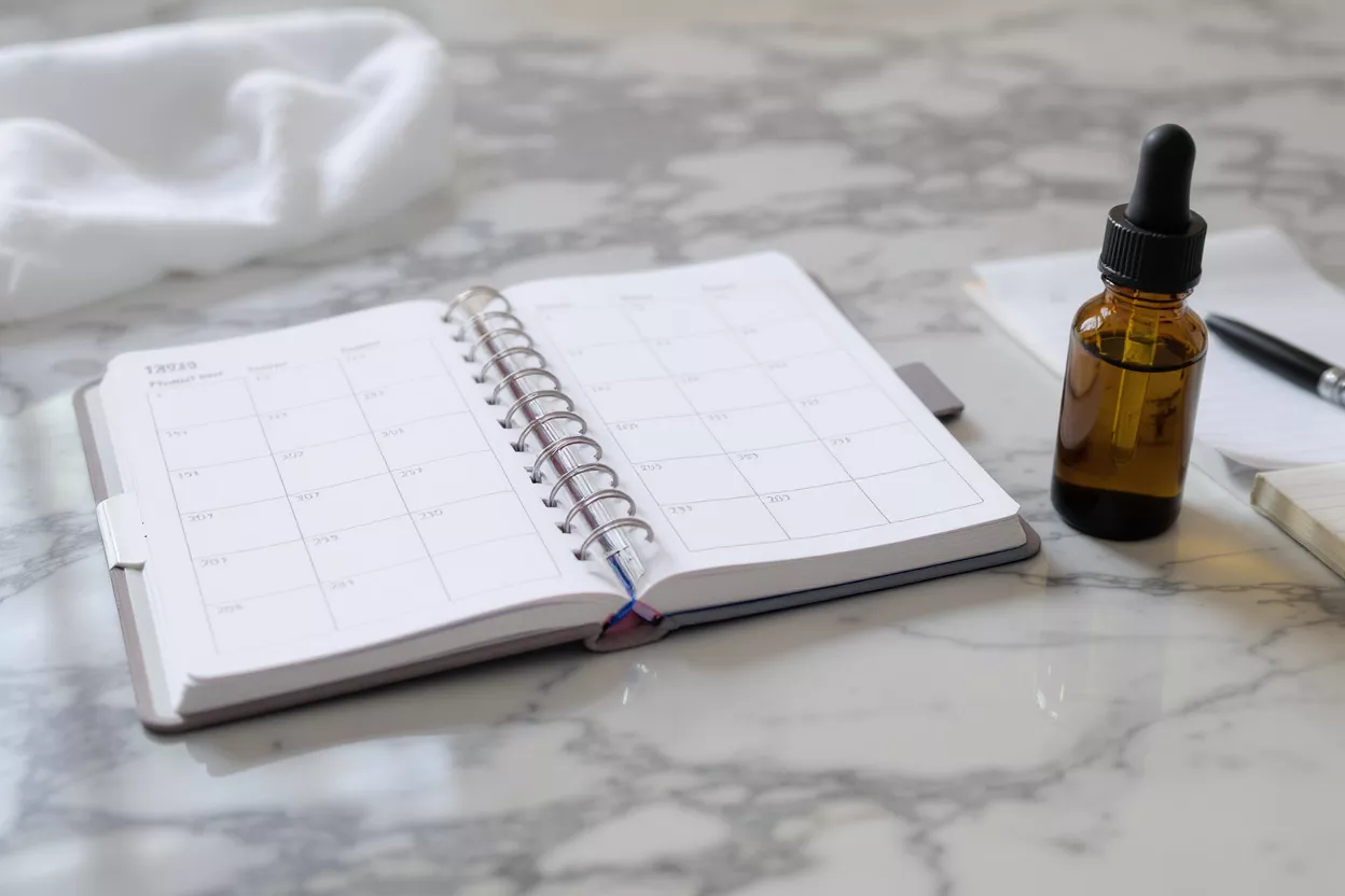 Open planner, amber dropper bottle, and pen on a white marble surface.