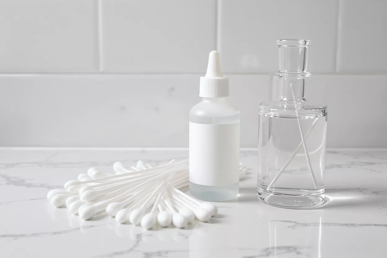White cotton swabs, a dropper bottle, and a clear glass bottle on a marble surface.