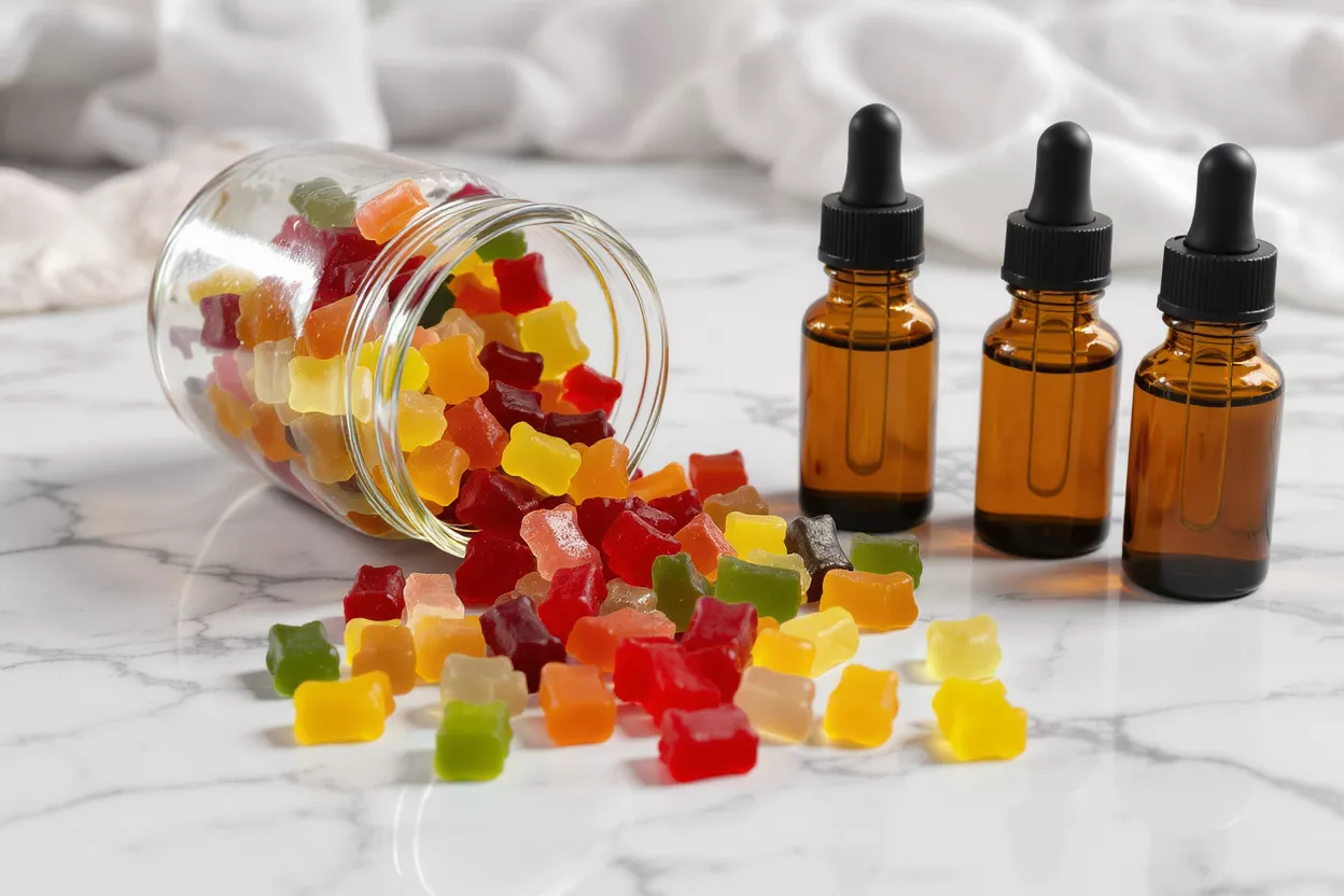Colorful CBD gummies spilled from a jar next to three CBD oil dropper bottles.