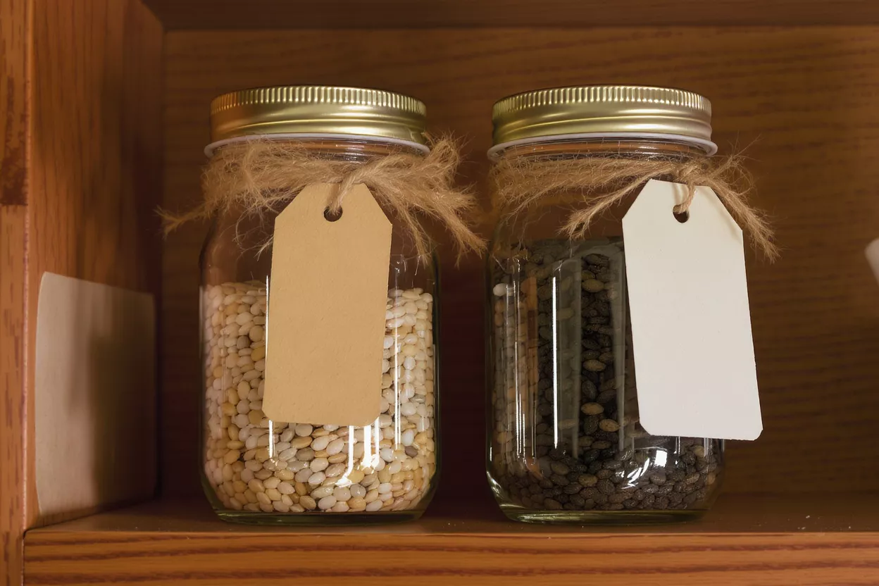 Two mason jars with seeds and blank tags on a wooden shelf.