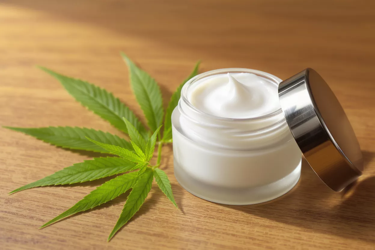 CBD Cream with Hemp Leaf A jar of white CBD cream with a hemp leaf on a wooden surface.