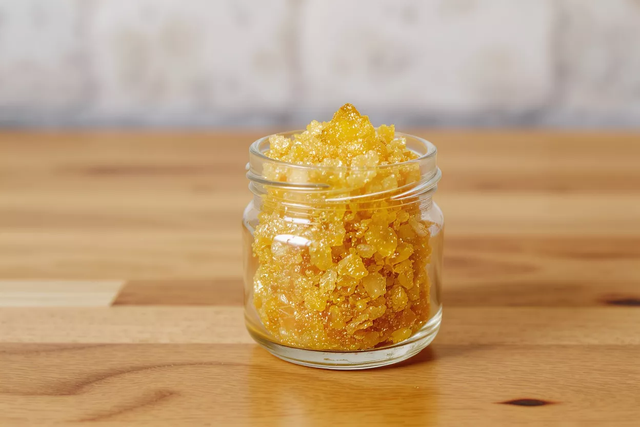 A small glass jar filled with golden, crystalline cannabis sugar wax concentrate on a wooden table.