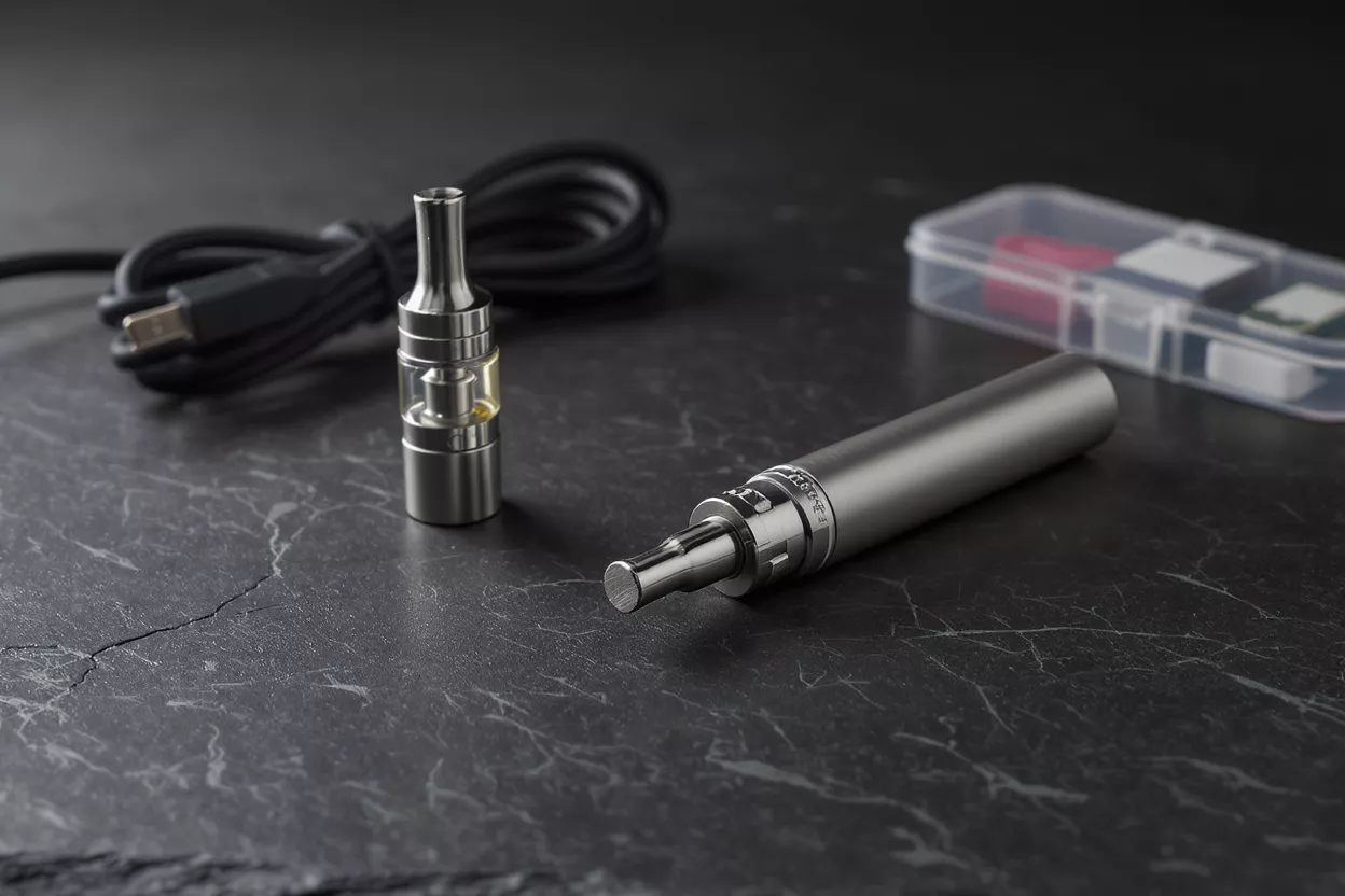 Vape pen, tank, USB charger, and clear case on a dark textured surface.