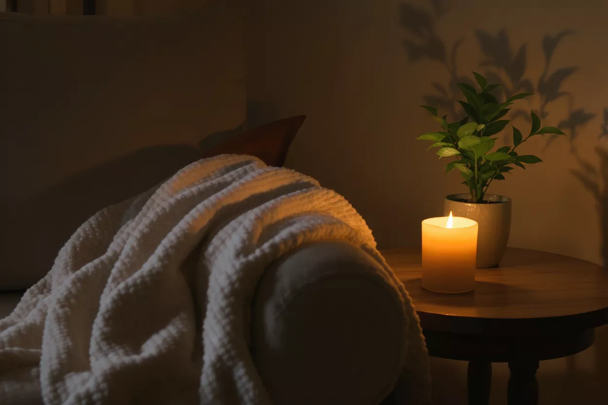 Warm candlelight illuminates a cozy blanket on a sofa next to a potted plant.