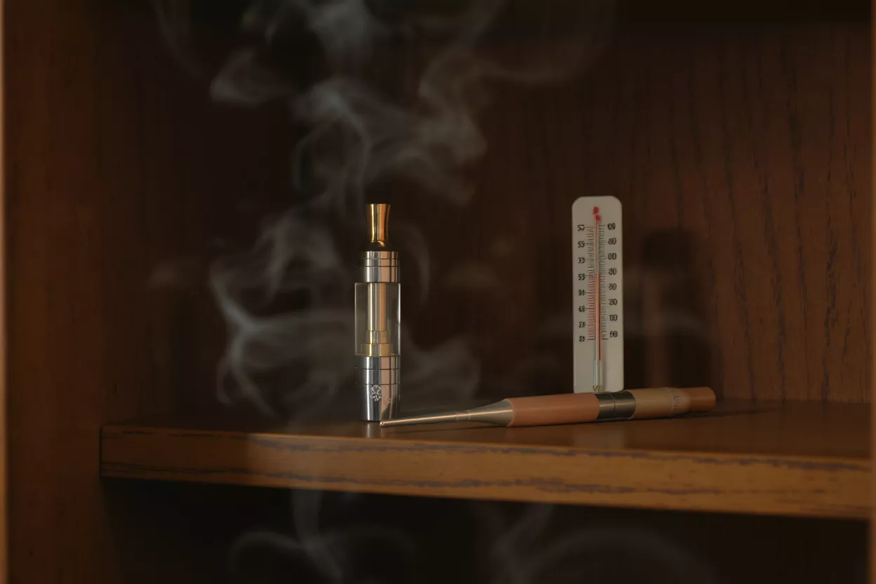 Dispositivo de vapeo, termómetro y lápiz óptico en un estante de madera con humo.