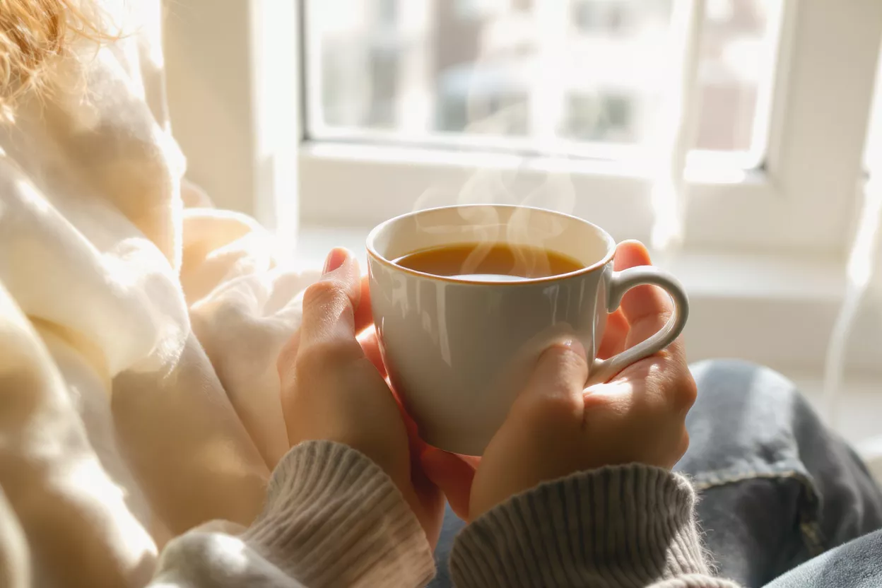 Café fumant à la lumière du matin Mains tenant une tasse à café blanche fumante près d'une fenêtre ensoleillée.