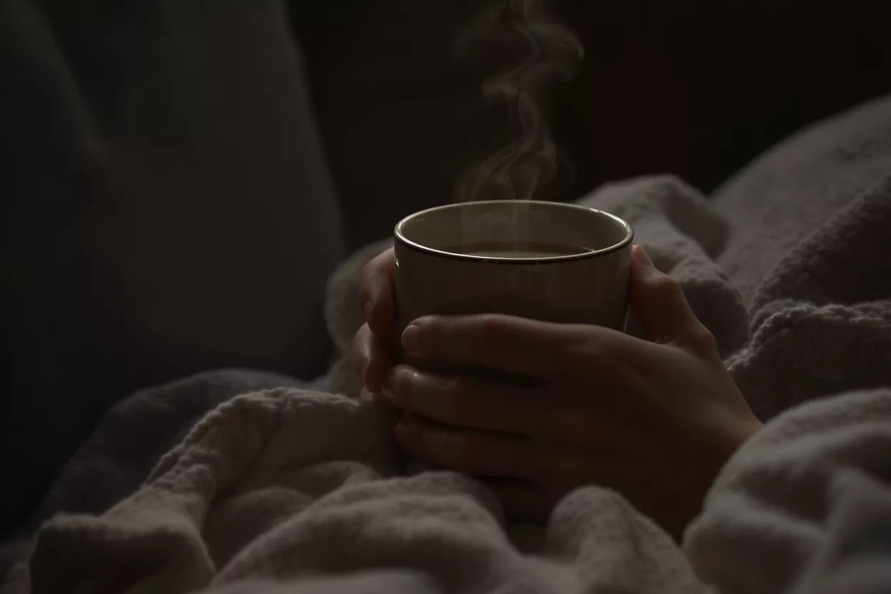 Hands holding a steaming mug, wrapped in a cozy blanket in dim light.