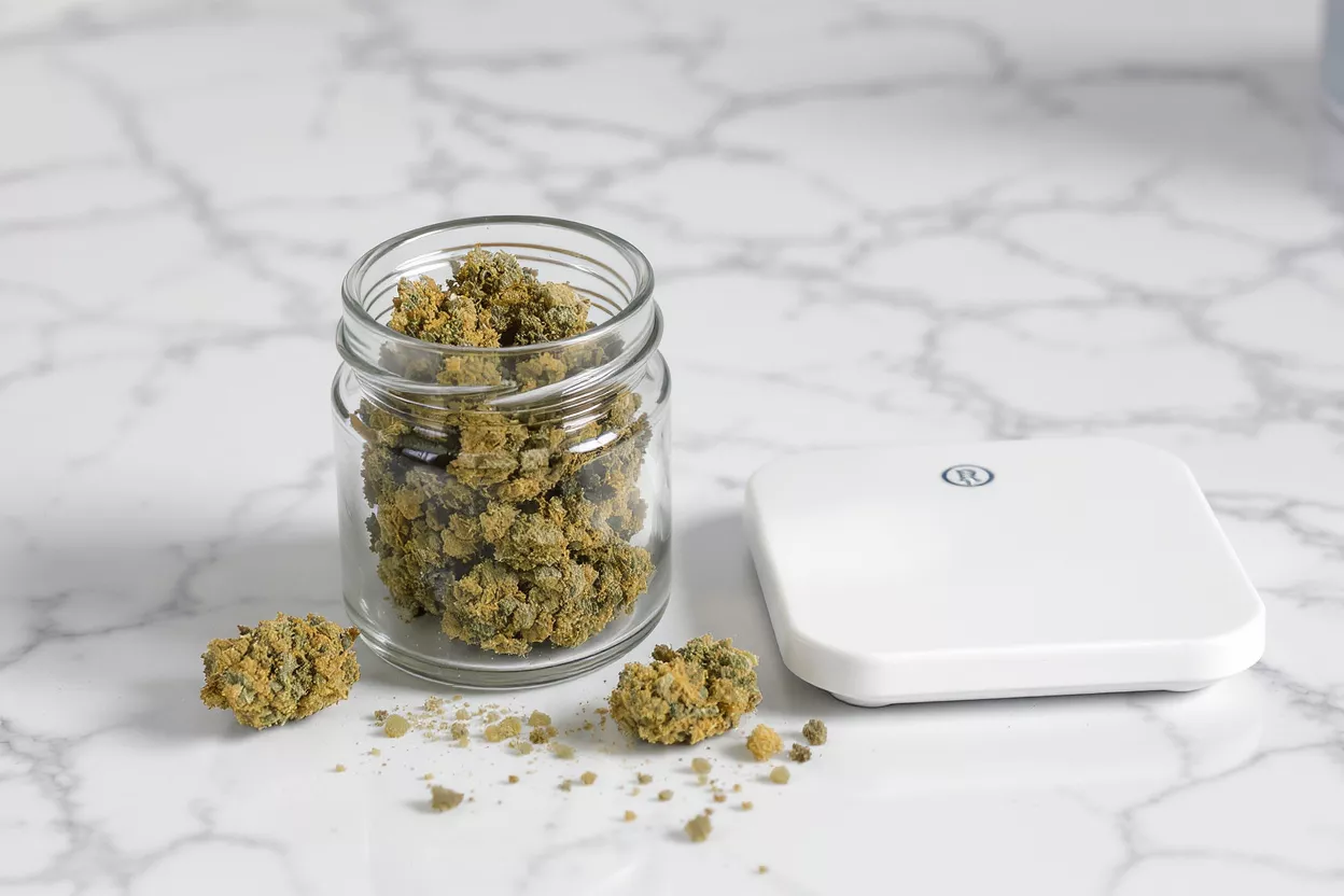 A glass jar filled with cannabis buds next to a white digital scale.