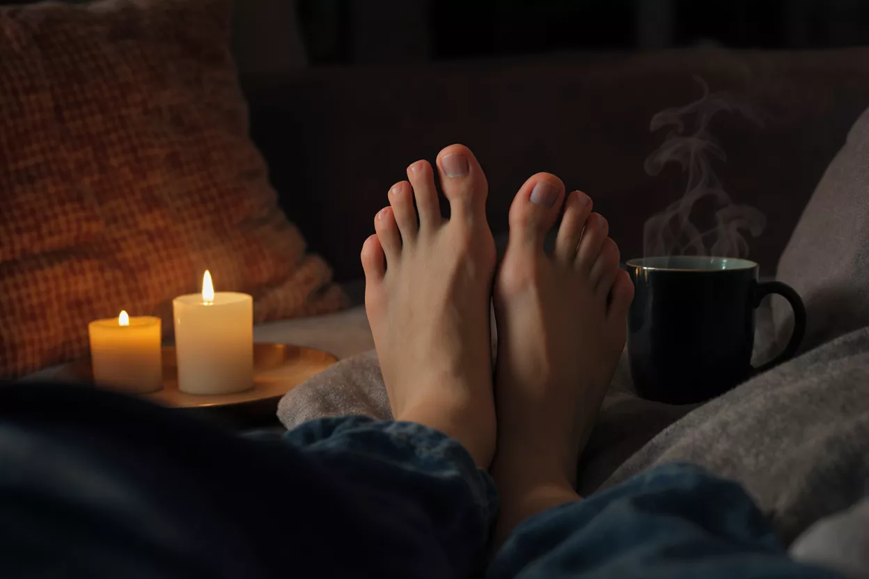 Feet relaxing on a couch with lit candles and a steaming mug.