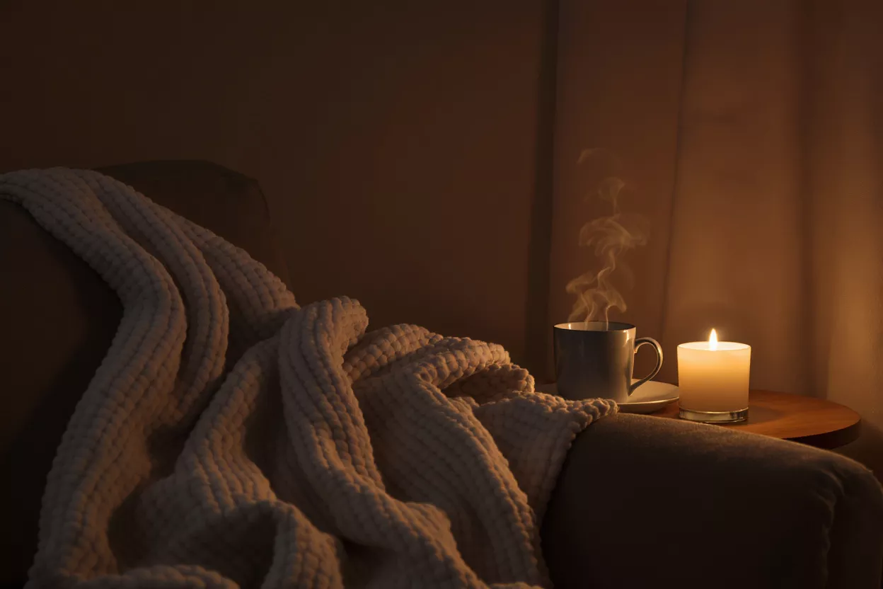 Steaming coffee mug, lit candle, and cozy blanket on an armchair in dim light.