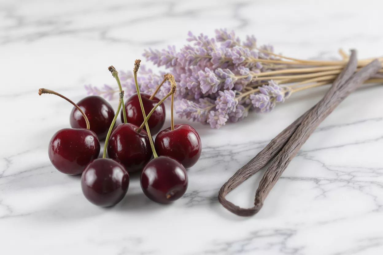 Dark red cherries, purple lavender, and vanilla beans on a white marble surface.