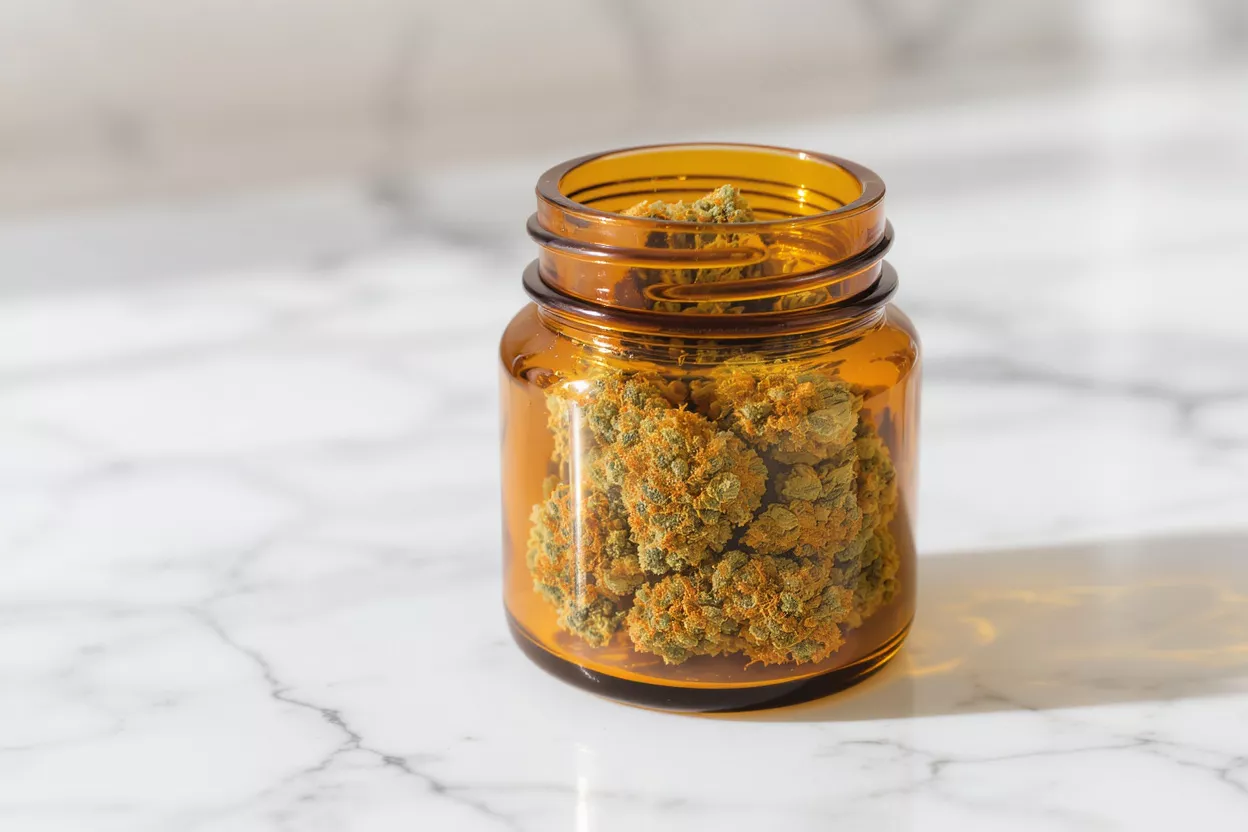 Close-up of vibrant cannabis buds in an amber glass jar on a white marble surface.
