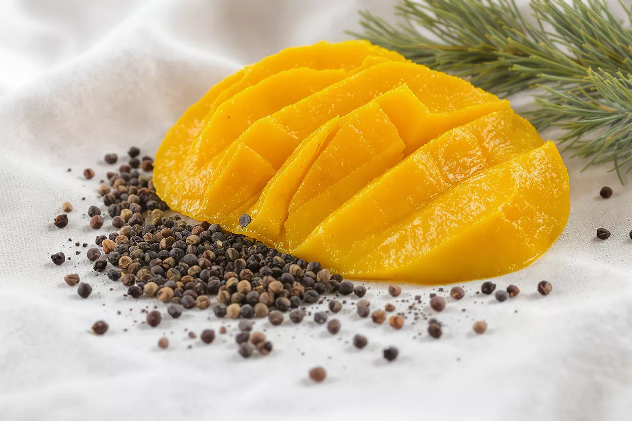 Bright yellow sliced mango with black peppercorns and green pine needles on white fabric.
