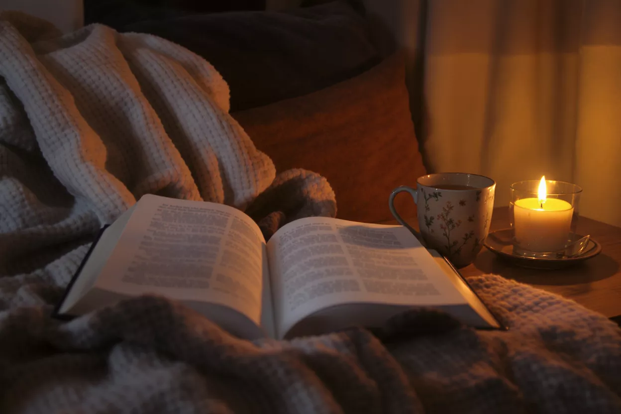 Open book, tea, and lit candle on a blanket in a cozy, dim room.