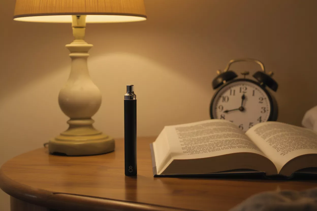 Black vape pen on a wooden bedside table with a lamp and open book.